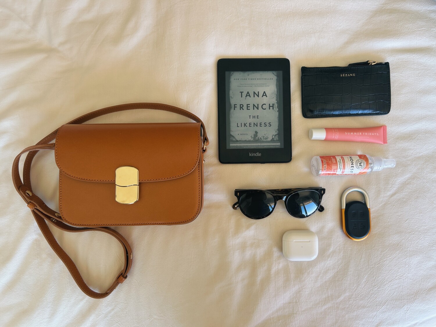 Sézane's Mini Milo bag next to a Kindle, sunglasses, card holder, lip gloss, and AirPods