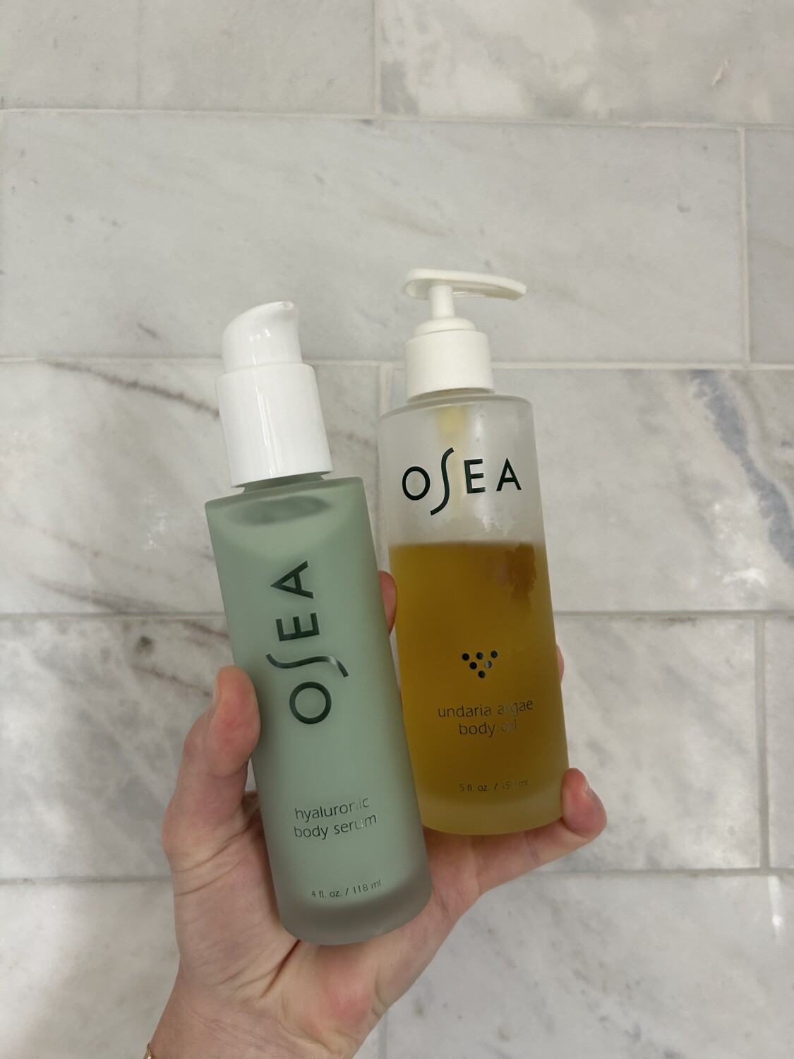 Osea body oil and body serum in front of a marble shower wall