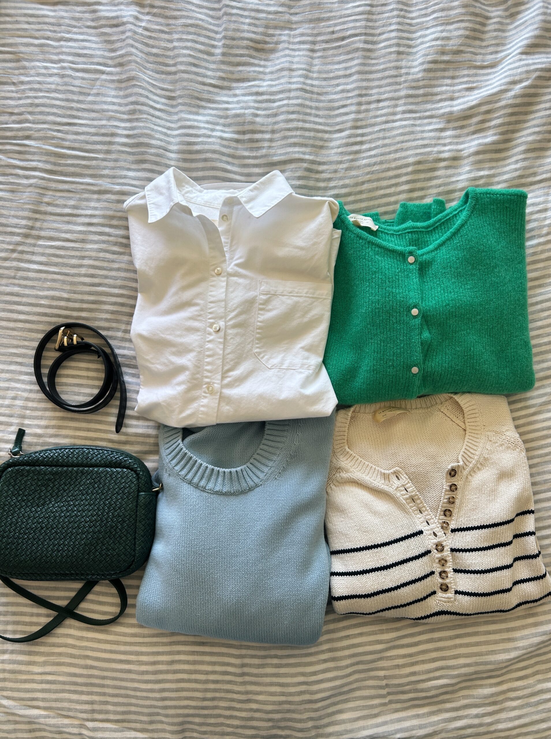 three sweaters, a button up shirt, a green purse, and a black belt laid out on a bed for a fall packing list for europe